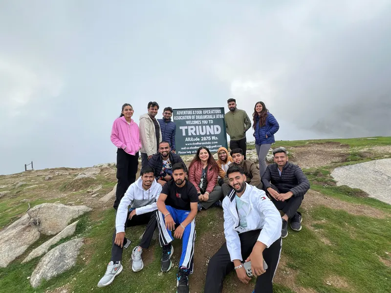 A group seated and standing beside the Triund sign on a hillside